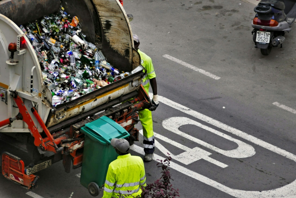 Photographie de deux ripeurs.euses derrière un camion poubelle